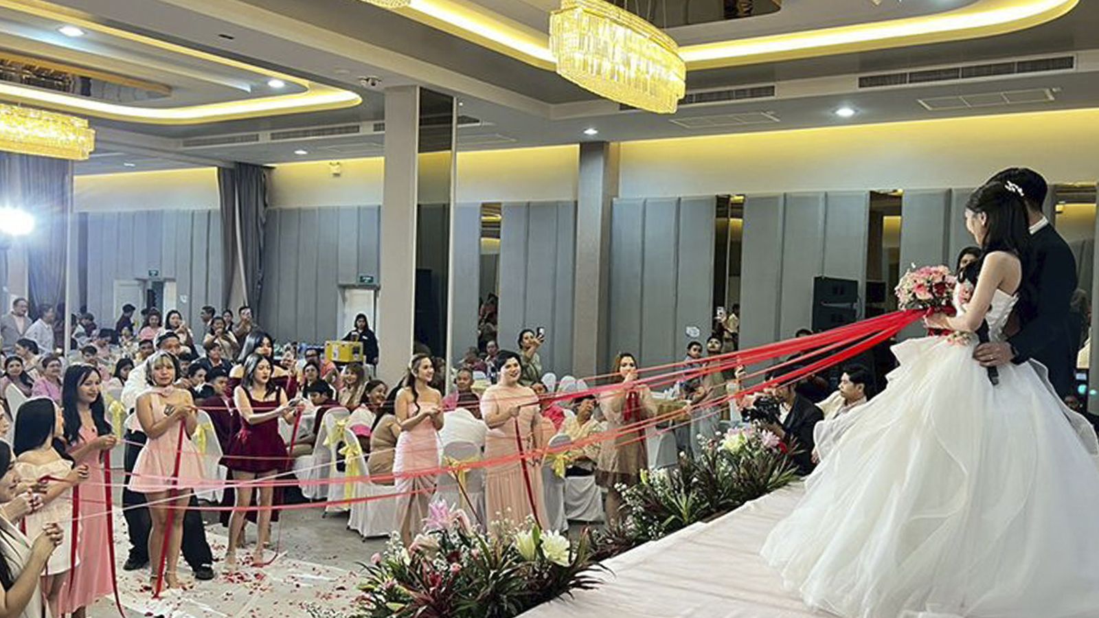 Bride and groom on stage as guests hold red ribbons across the ballroom